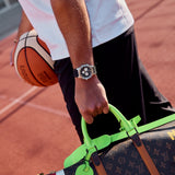 A close-up lifestyle shot of a person on a basketball court holding a Louis Vuitton monogram keepall bag with bright neon green handles and a matching tag, while wearing a Cornavin watch.