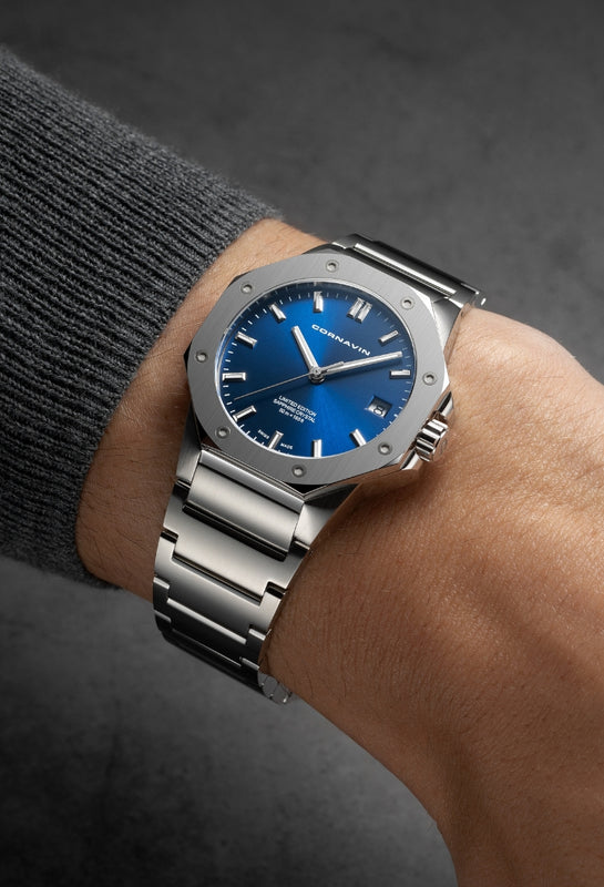 A wrist wearing a silver-toned Cornavin watch with a blue dial and silver bezel, shown against a dark gray textured background.